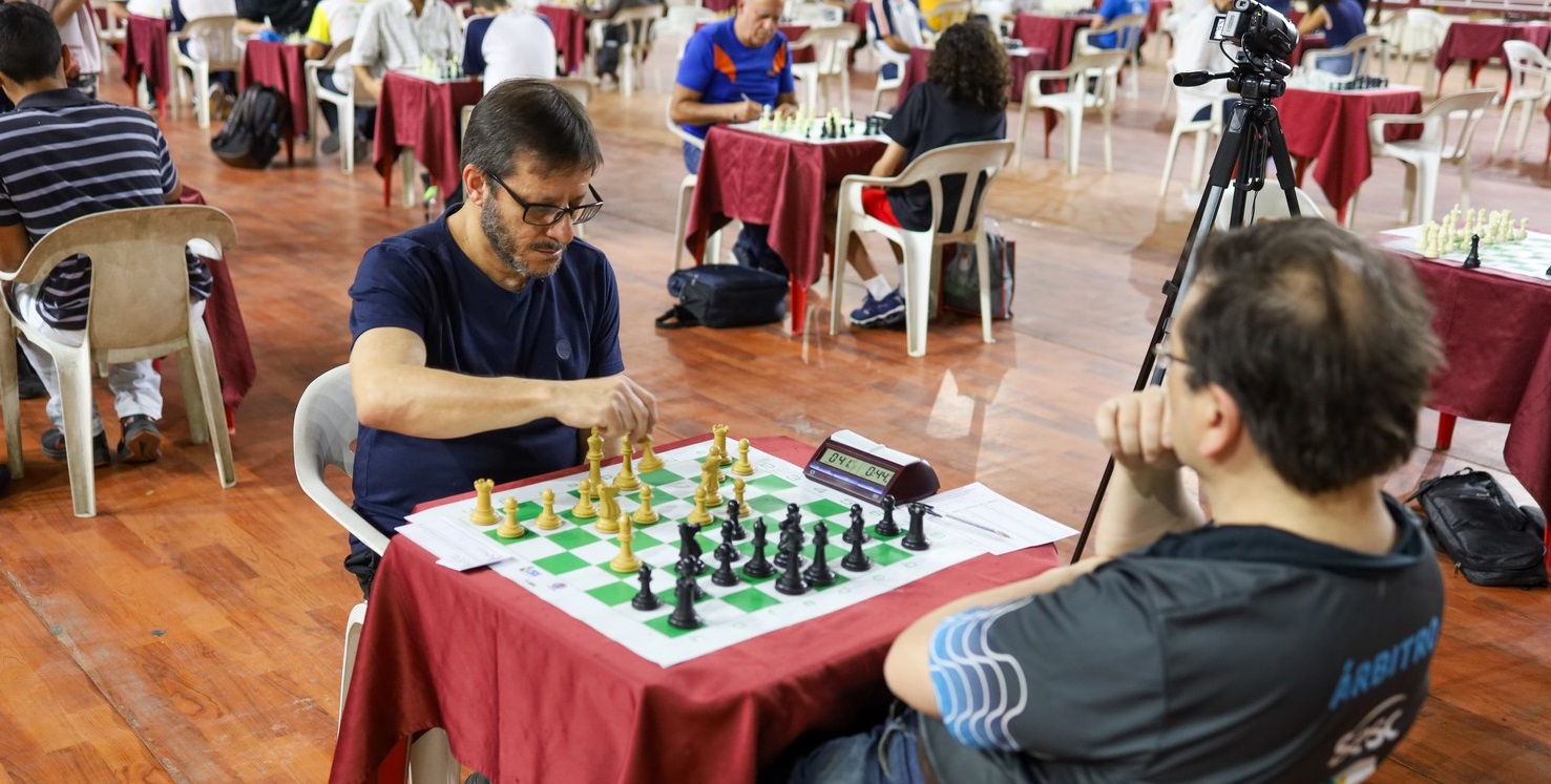 imagem mostra dois jogadores de xadrex em uma mesa. de costas há um homem não identificado, e de frente vemos o professor péricles